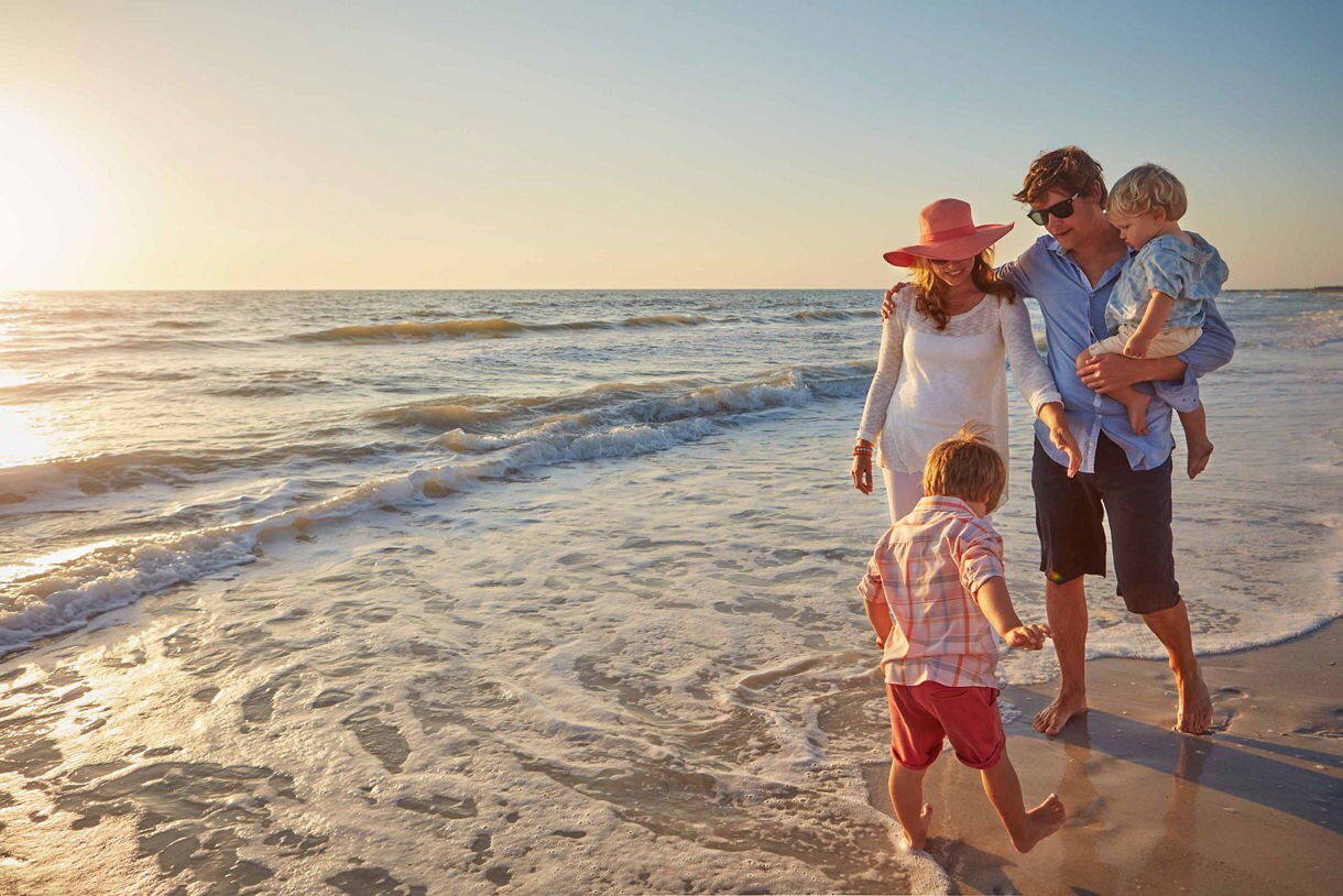 A family of four walks barefoot along the shoreline at sunset, with parents dressed in light beachwear and children playing in the gentle ocean waves.