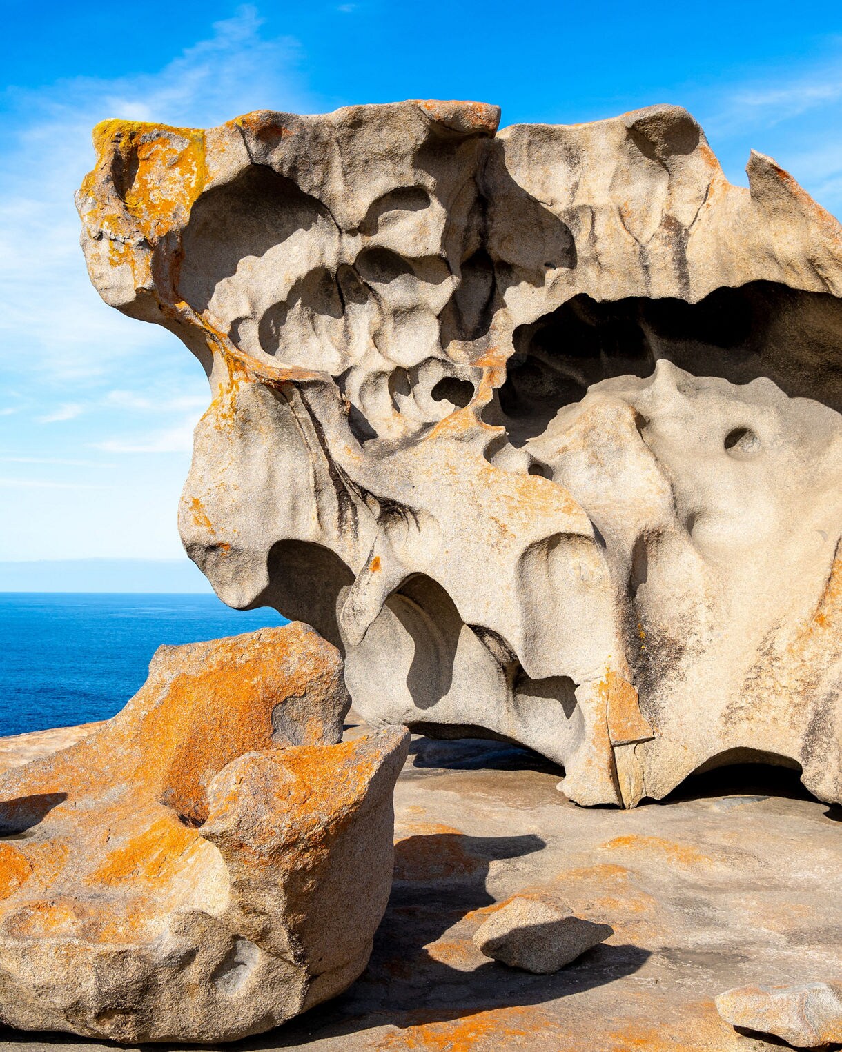 Weather-sculpted granite boulders with orange lichen overlooking deep blue sea under a bright sky.