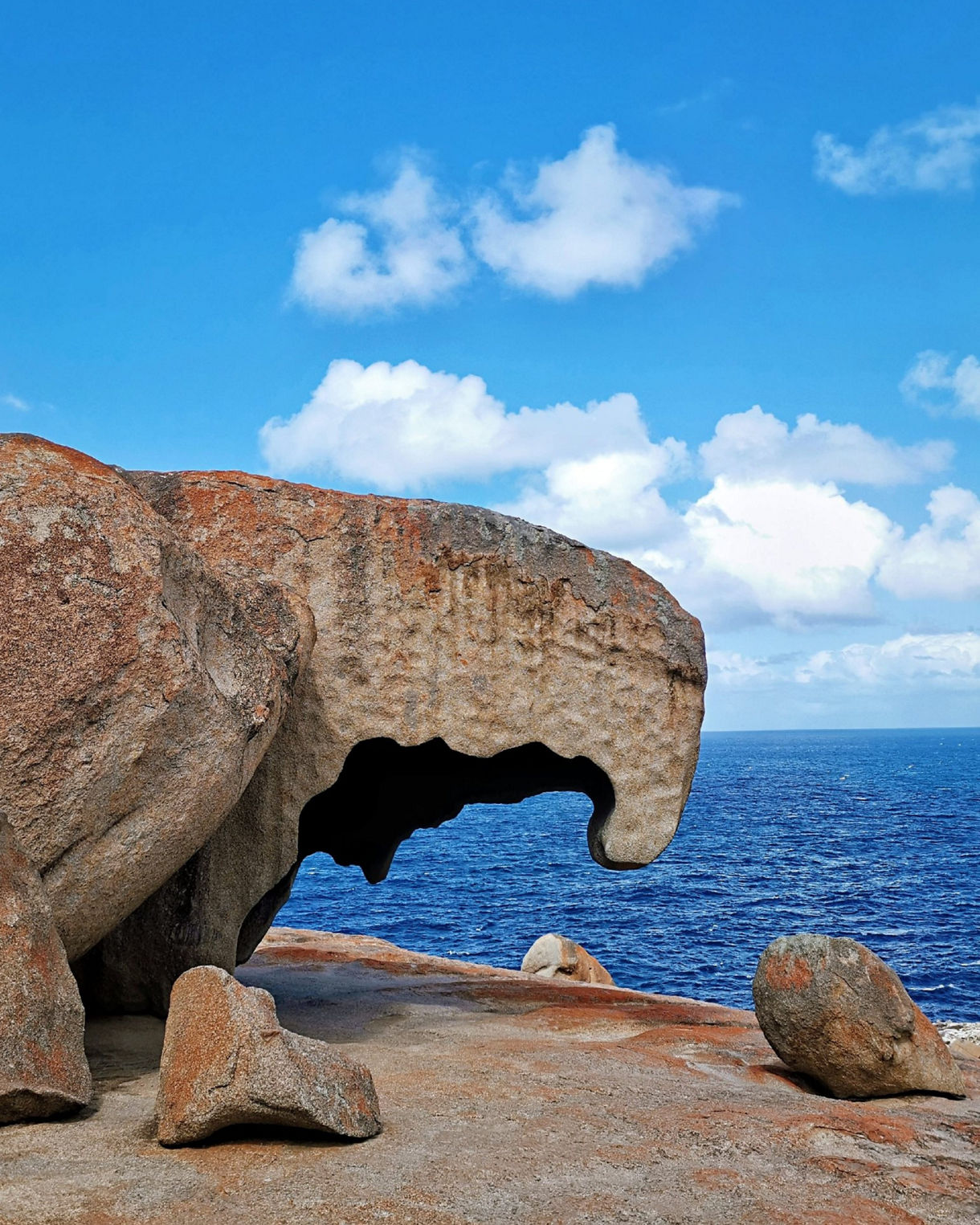 Curved granite formation with orange lichen perched above deep blue ocean under a bright sky with scattered clouds.