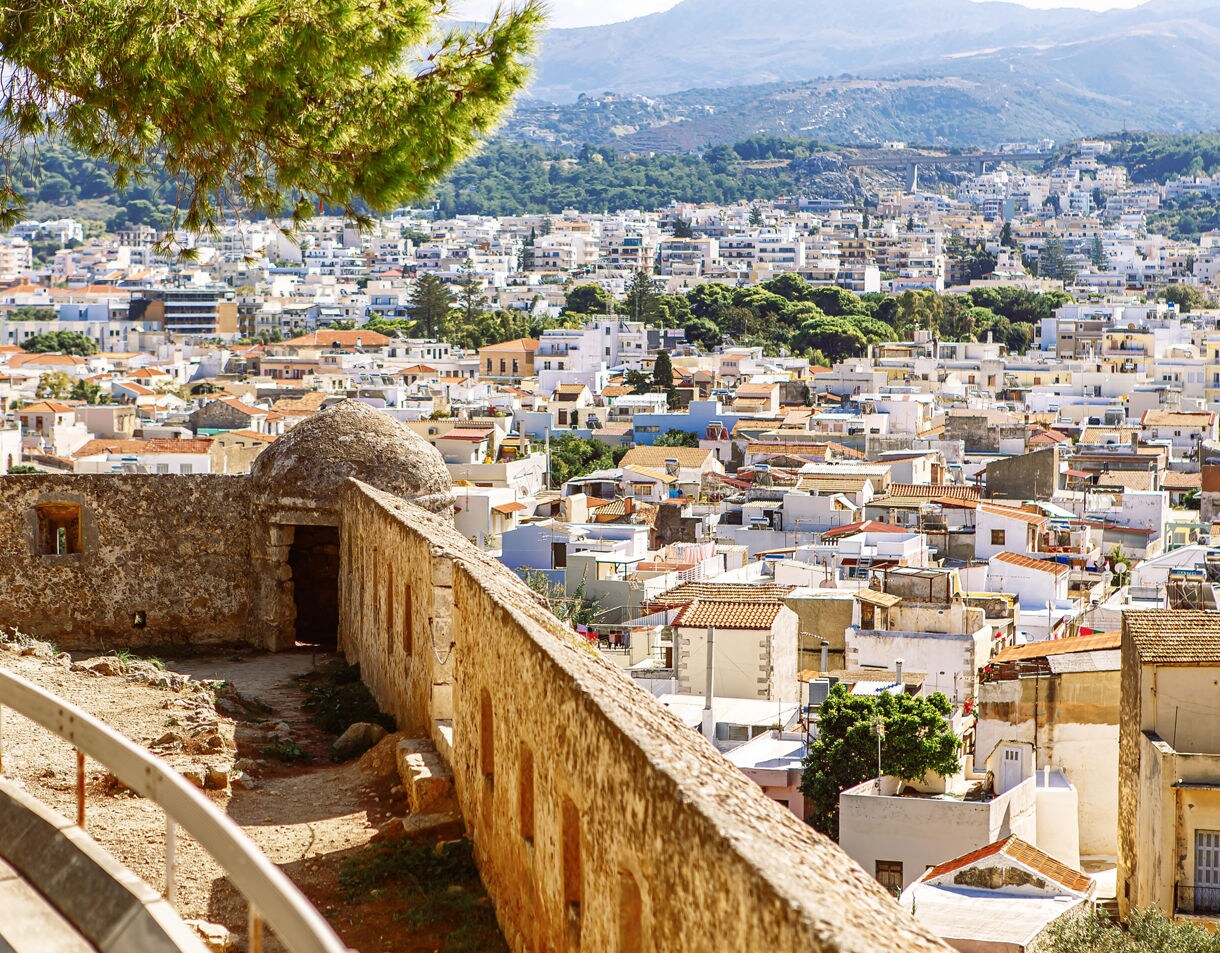 Elevated view from the Venetian Fortezza in Rethymnon, Crete, showing the stone walls overlooking a sea of whitewashed homes and terracotta roofs with mountains in the distance.