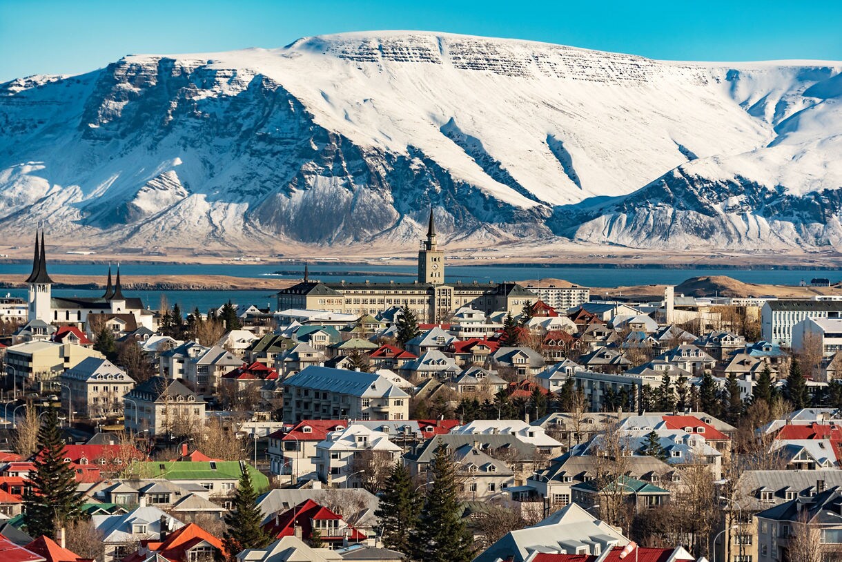Panoramic view of Reykjavik, Iceland, with colorful rooftops, church spires and a backdrop of towering snow-covered mountains near the coastline.