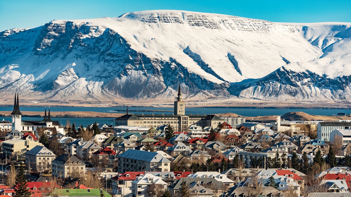 Panoramic view of Reykjavik, Iceland, with colorful rooftops, church spires and a backdrop of towering snow-covered mountains near the coastline.