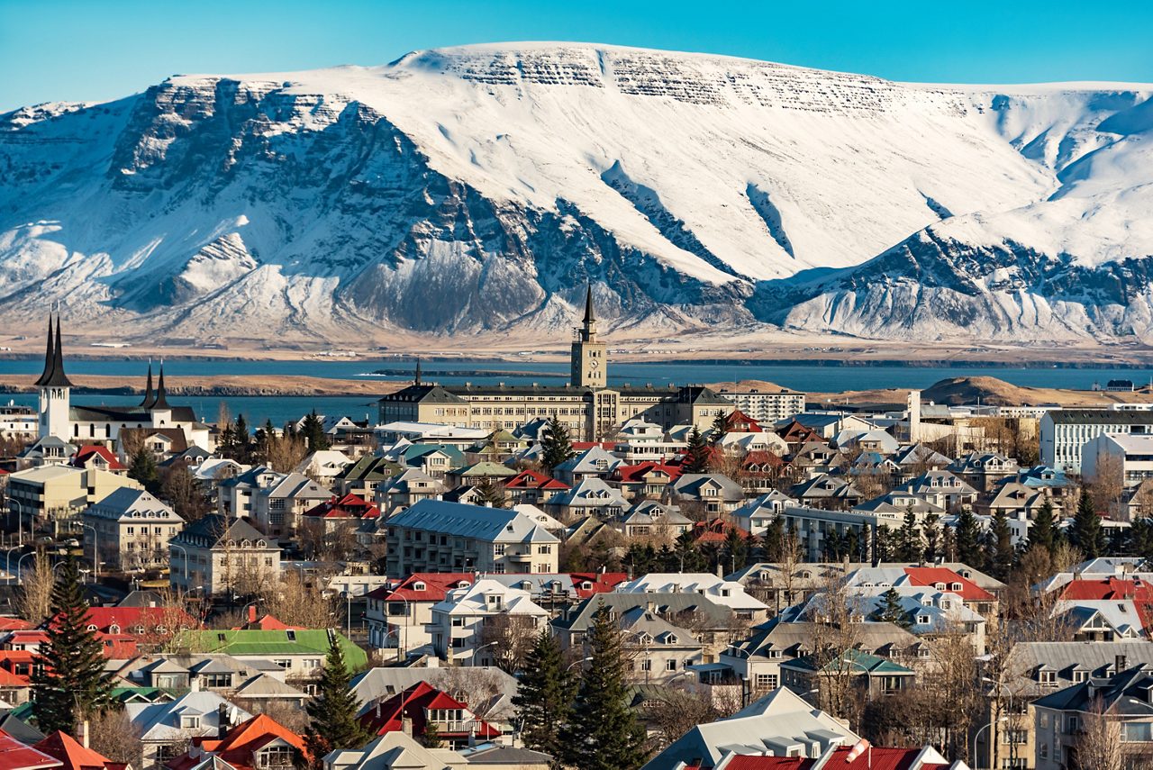 Panoramic view of Reykjavik, Iceland, with colorful rooftops, church spires and a backdrop of towering snow-covered mountains near the coastline.
