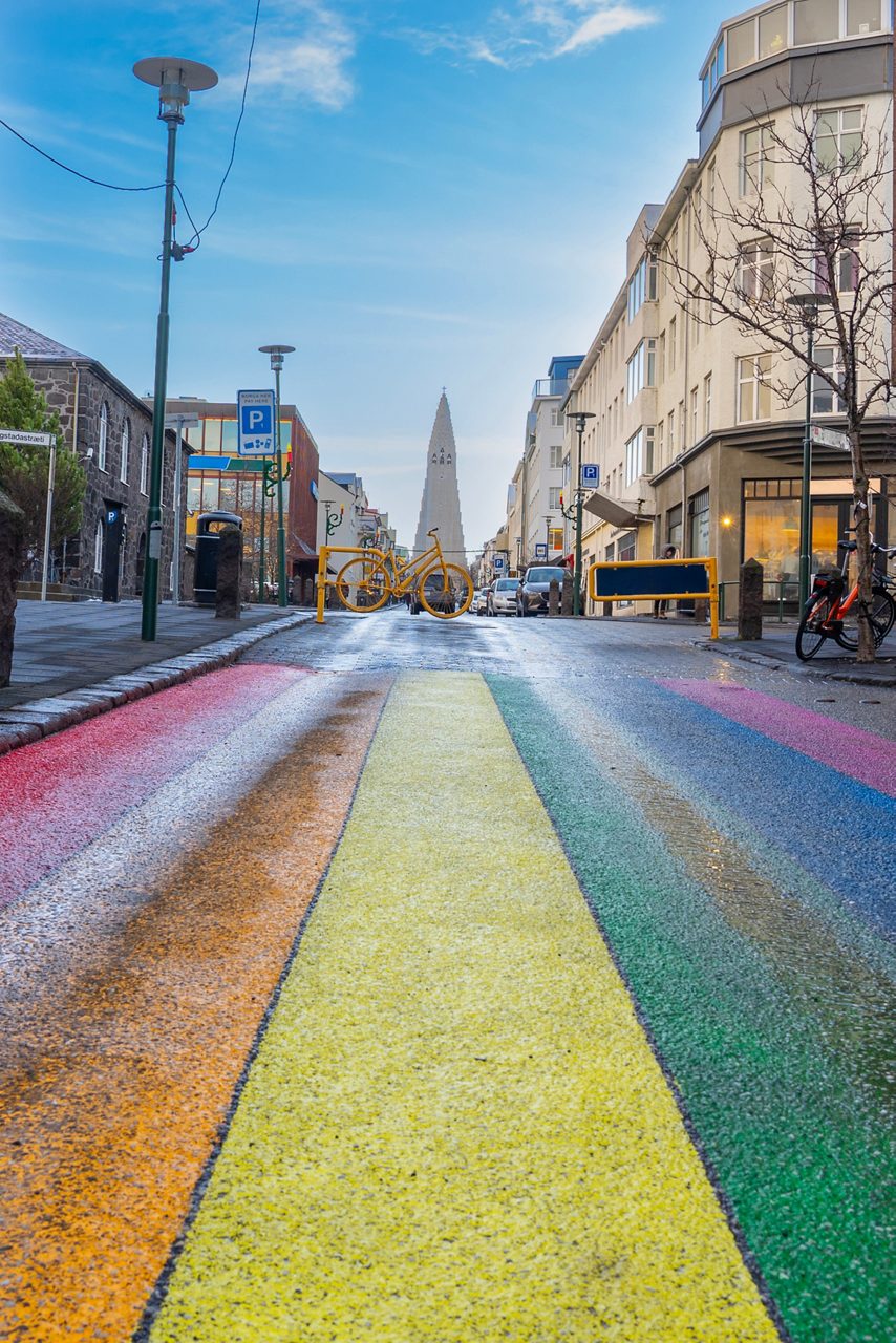Rainbow-painted street in Reykjavik, Iceland, with colorful stripes leading toward Hallgrímskirkja church tower, framed by city buildings under a clear blue sky.