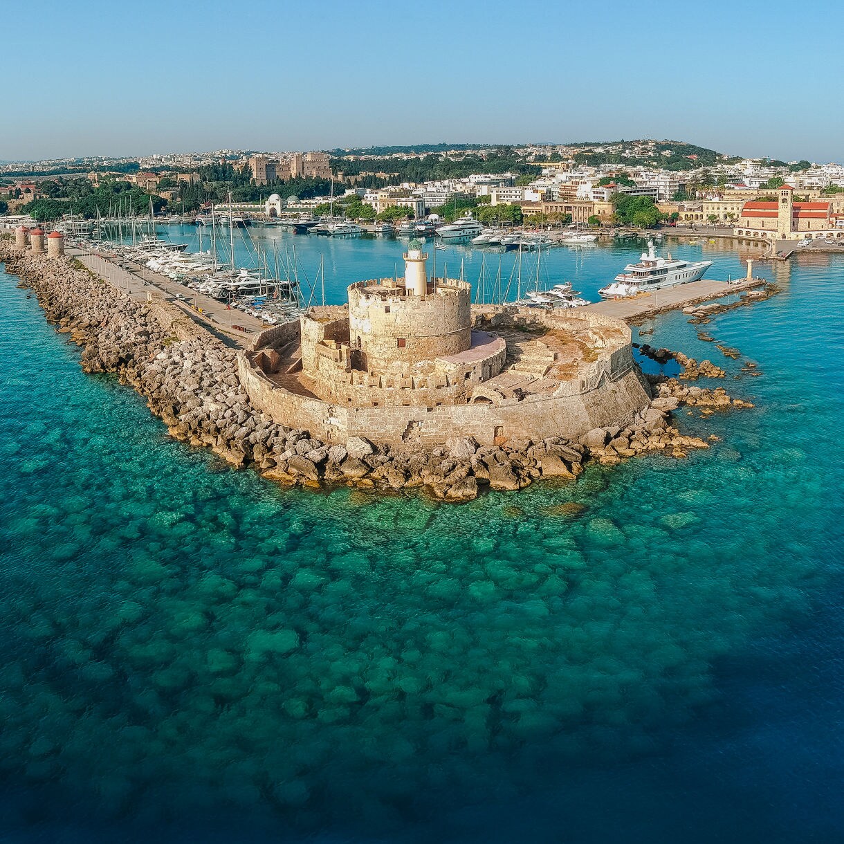 Aerial view of the historic harbor of Rhodes, Greece, featuring a stone fortress and lighthouse surrounded by turquoise water and moored yachts.