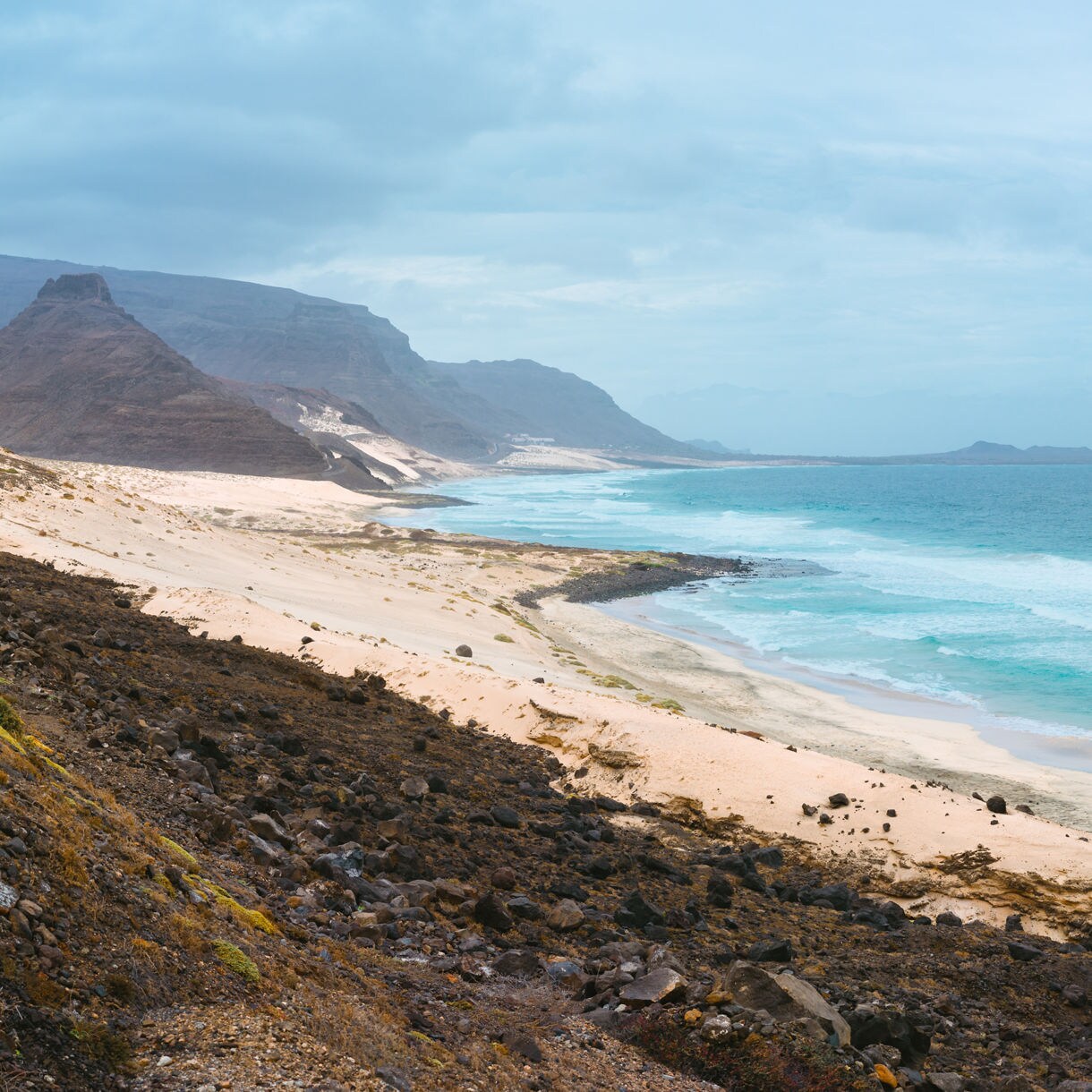 Panoramic view of a rugged coastal highway skirting a wide sandy beach with turquoise waves, dark volcanic hills rising to the left and distant headlands under a muted, cloudy sky.