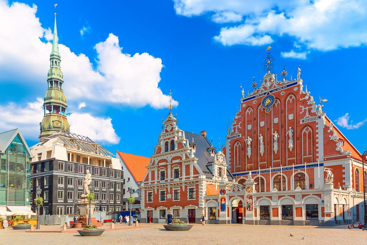 View of Riga’s Town Hall Square in Latvia, featuring the House of the Blackheads with red brick façade and statues, alongside St. Peter’s Church tower rising above the skyline under a bright blue sky.