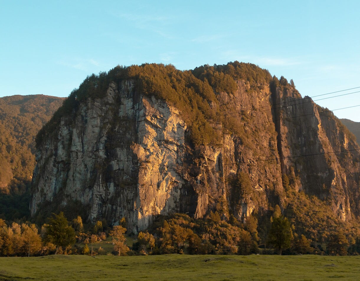 Sheer rock cliffs bathed in warm sunset light, surrounded by forested hills and grassy fields in Río Simpson National Reserve.