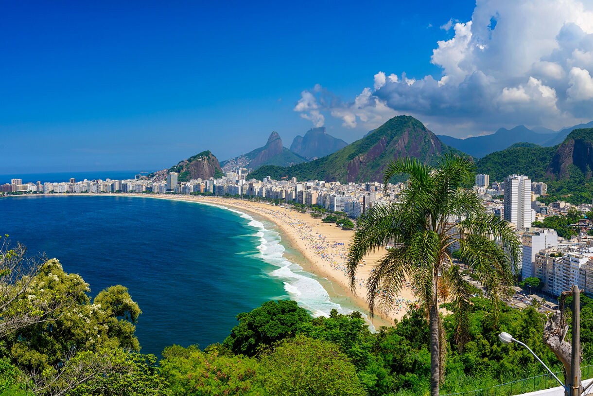 Panoramic view of Rio de Janeiro showing Copacabana Beach, blue ocean waves, dense city buildings, lush green mountains and a Brazilian flag waving in the foreground.
