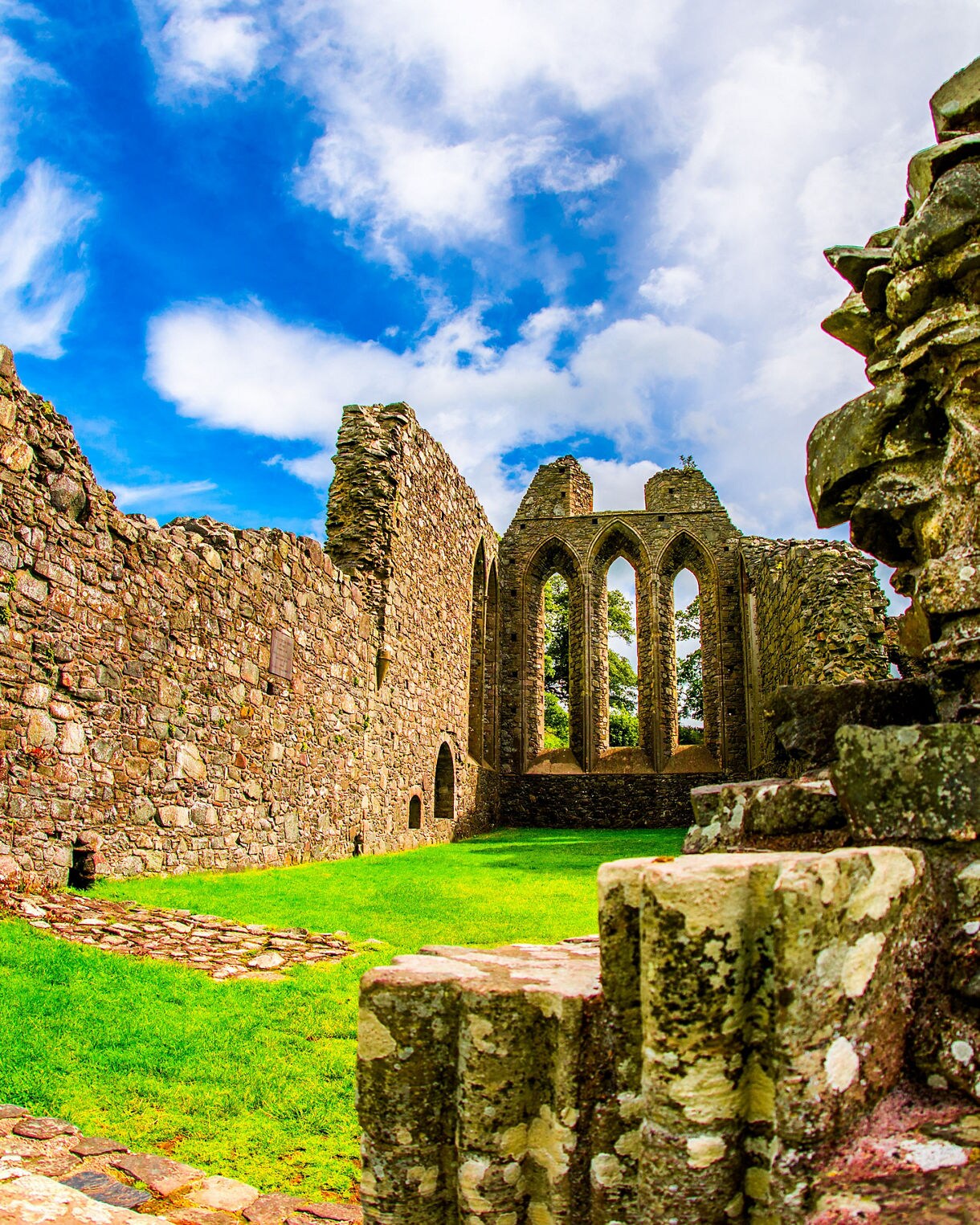 Stone ruins of Inch Abbey in Northern Ireland with arched Gothic windows, crumbling walls and lush green grass under a bright blue sky.