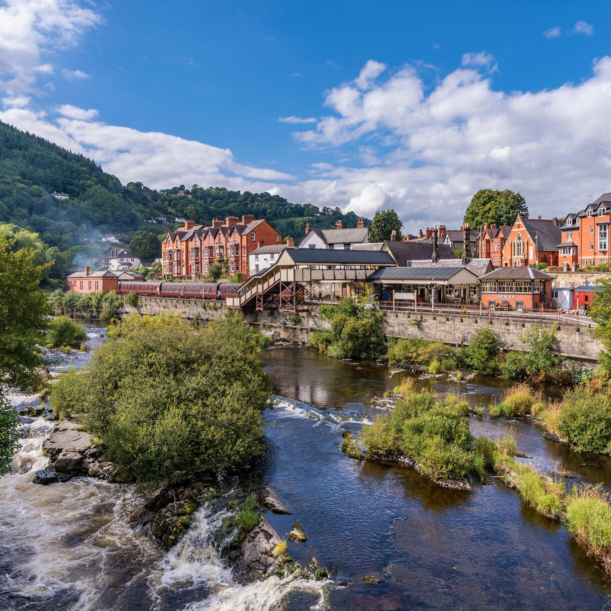 View of Llangollen, Wales, showing a riverside town with red-brick architecture, a railway station and surrounding green hills.