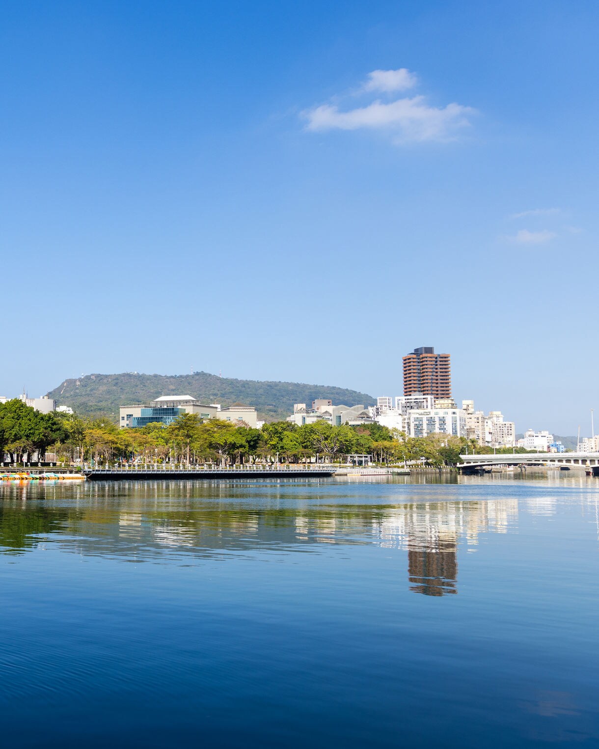 Peaceful view of Kaohsiung’s Love River with reflections of modern buildings, green trees and distant hills under a bright blue sky.