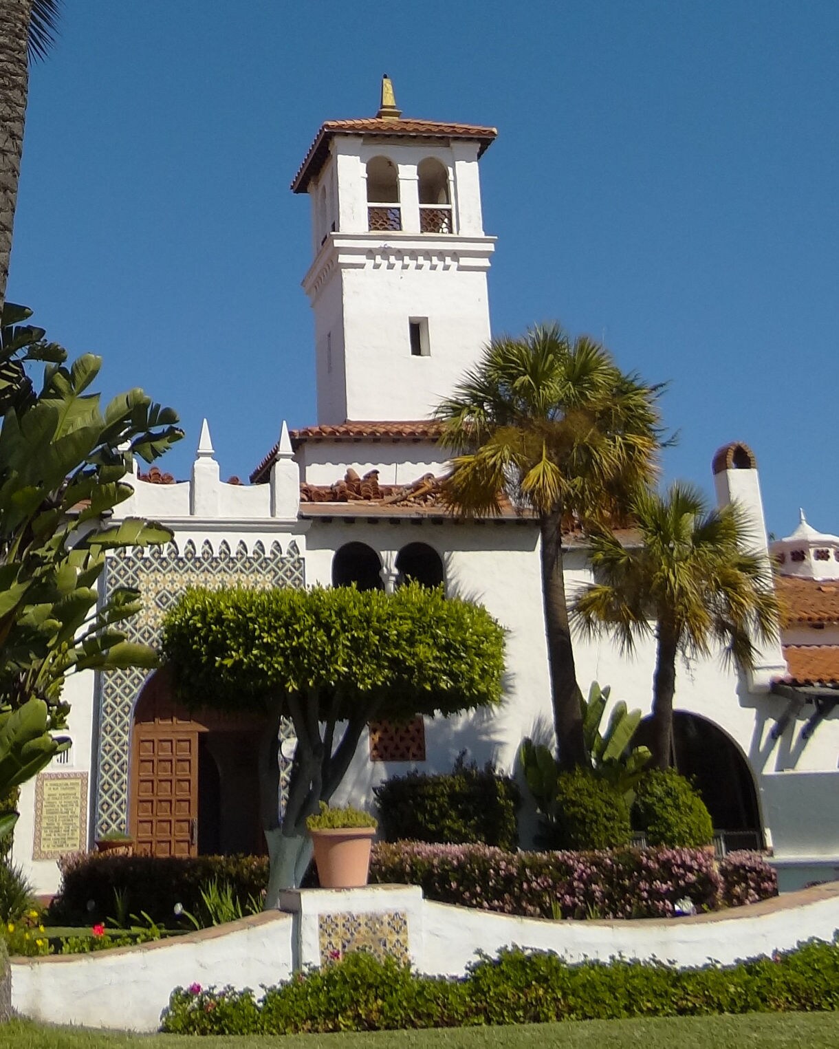 White stucco building with red-tiled roof, arched windows and tower at Riviera del Pacífico in Ensenada, surrounded by palms and gardens.