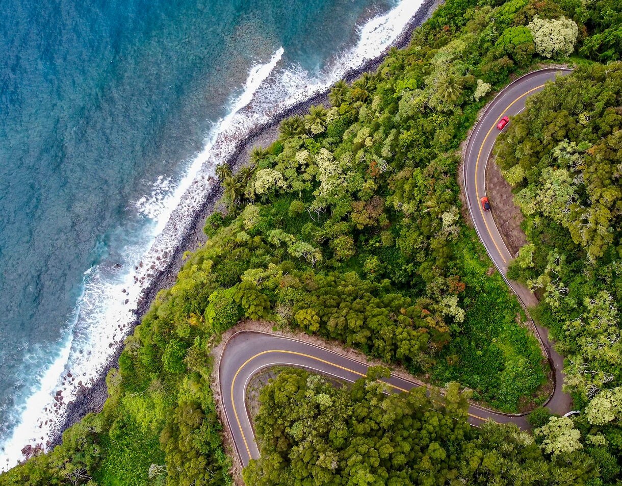 Aerial view of the winding Road to Hana bordered by dense green jungle and the blue ocean shoreline.