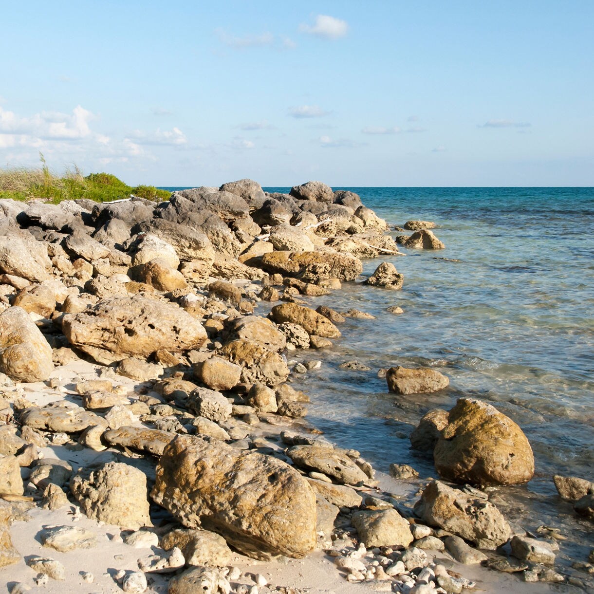 Rocky shoreline at Taino Beach with sunlit boulders scattered along the sand beside calm turquoise water.