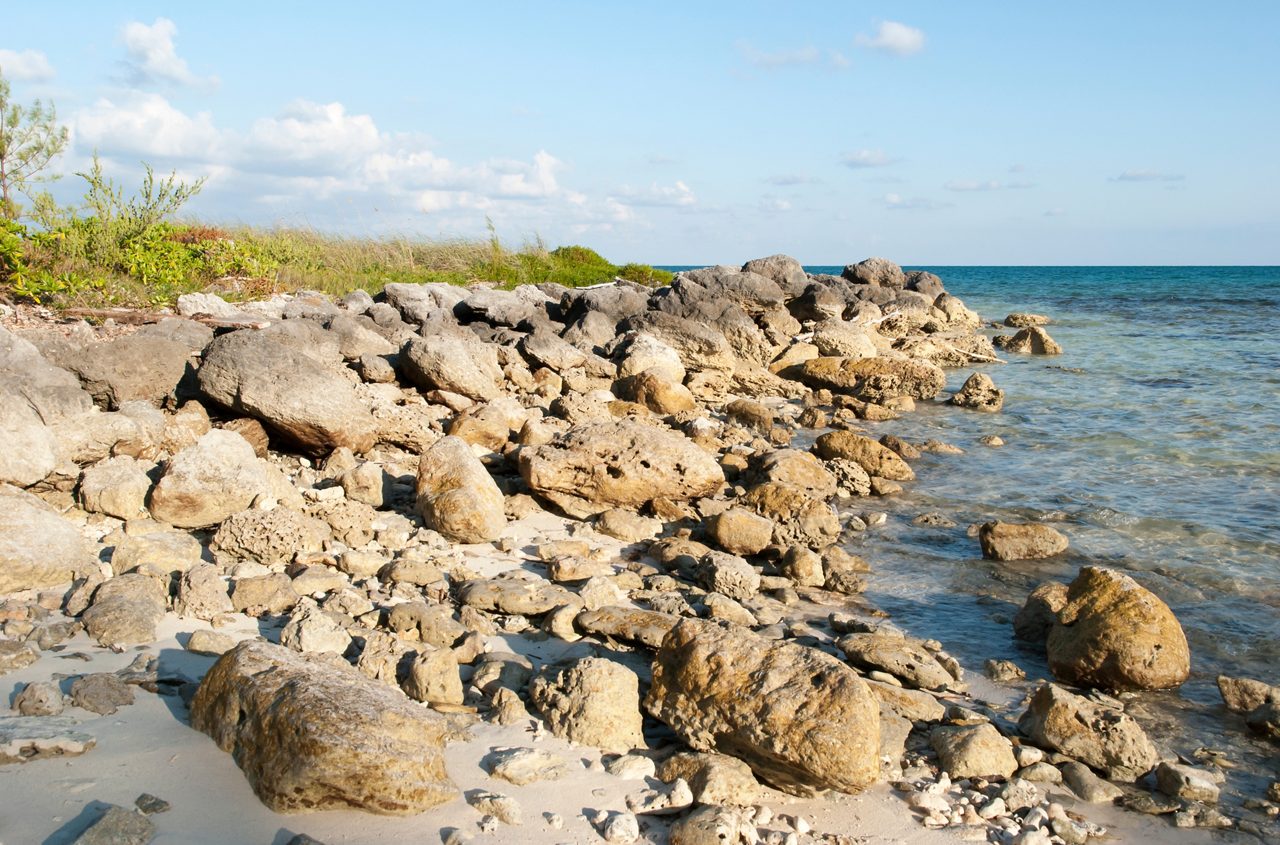 Rocky shoreline at Taino Beach with sunlit boulders scattered along the sand beside calm turquoise water.