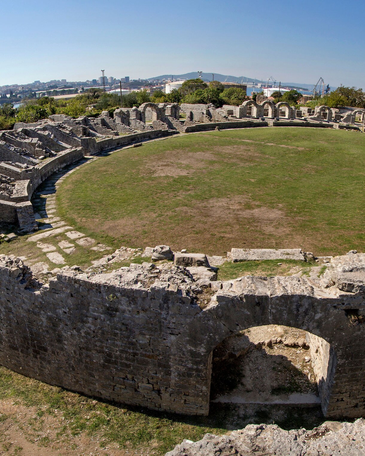 Wide view of a ruined Roman amphitheater with a grassy central arena, crumbling stone seating and walls surrounding it, under clear blue skies near Split, Croatia.