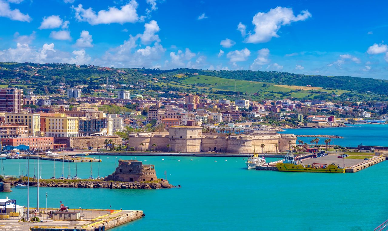 A panoramic aerial view of the port of Civitavecchia, Rome's cruise port, featuring vivid turquoise water, an ancient stone fortress, colorful waterfront buildings, docked vessels and rolling green hills in the background under a bright blue sky.