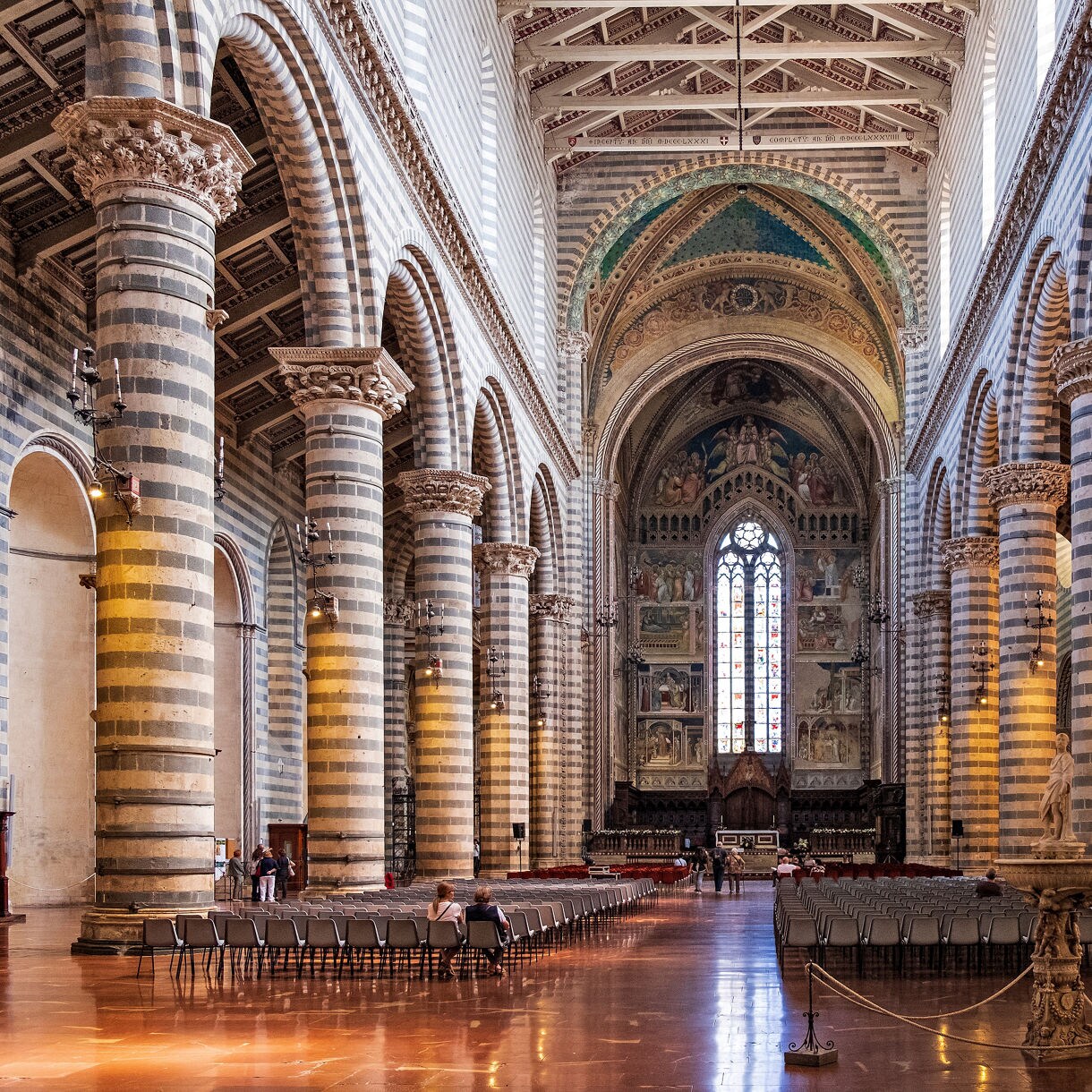 Interior of Orvieto Cathedral in Italy with striped stone columns, vaulted ceilings, and a stained-glass window above the ornate altar.