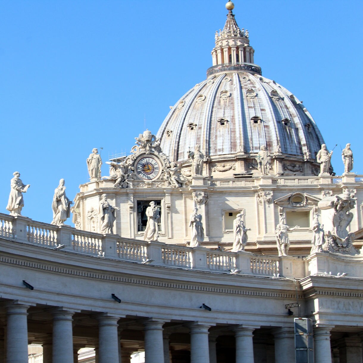 Close-up of St. Peter’s Basilica in Vatican City, showing the dome, clock, and statues atop the colonnade under a clear blue sky.