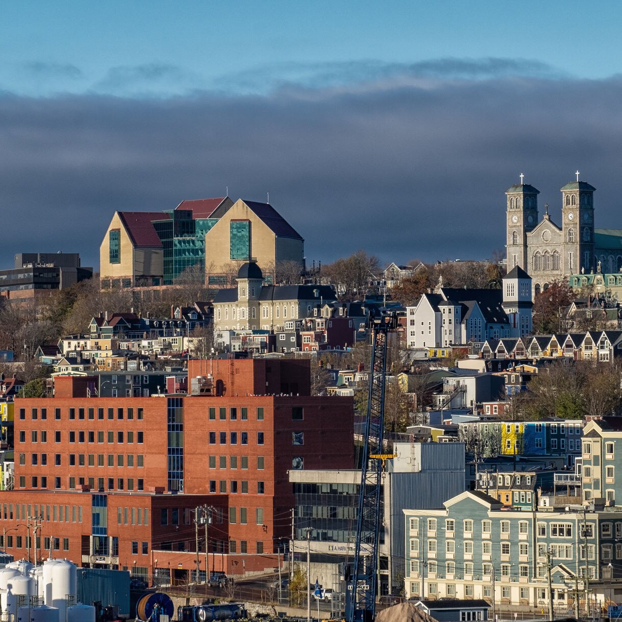 A panoramic view of St. John’s, Newfoundland, showing colorful row houses, modern buildings, large red-brick structures and prominent landmarks like the Rooms museum and the twin-tower basilica under a clear blue sky.