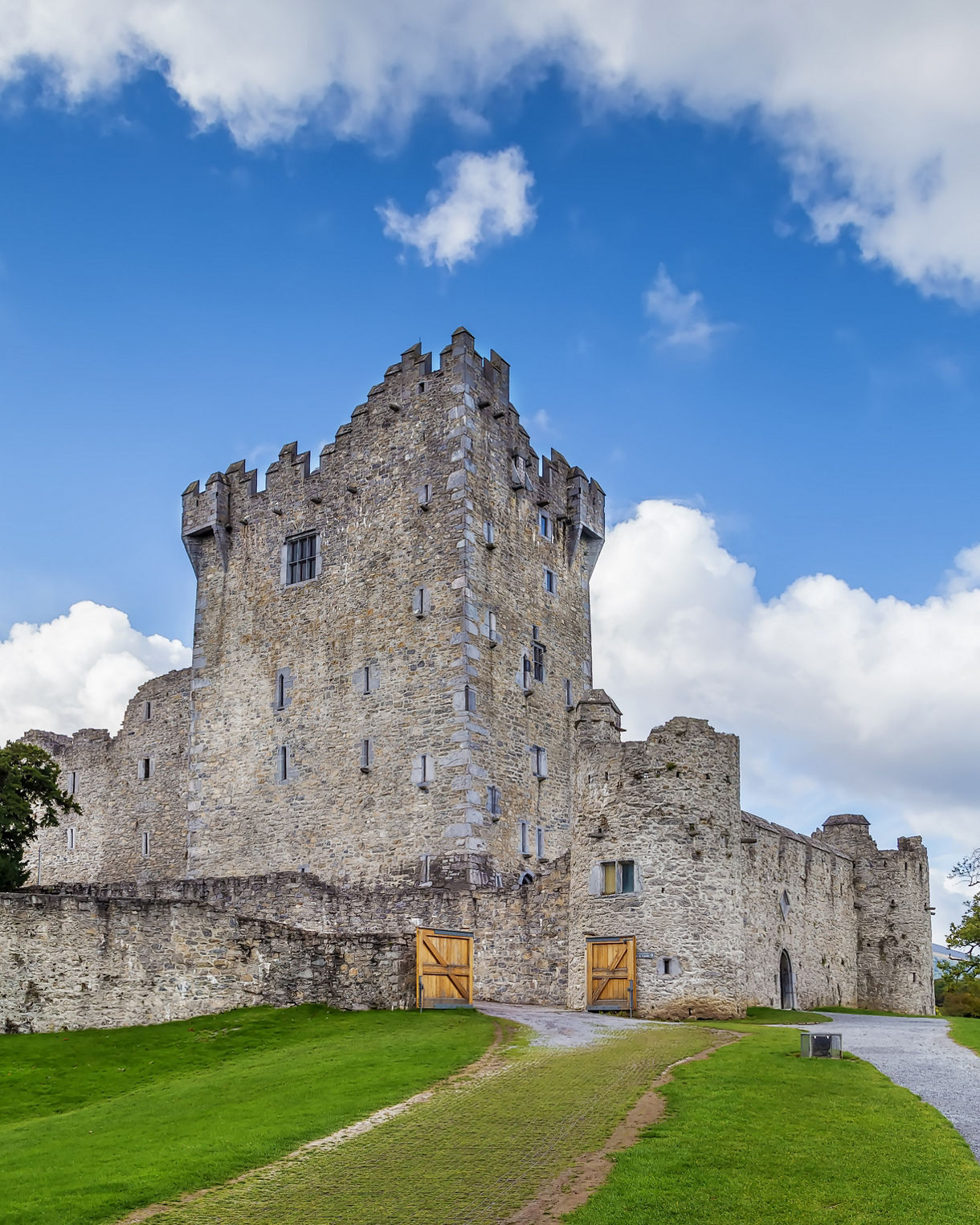 Ross Castle in Killarney National Park, Ireland, a tall stone tower with battlements and surrounding walls, set against a bright blue sky with scattered clouds.