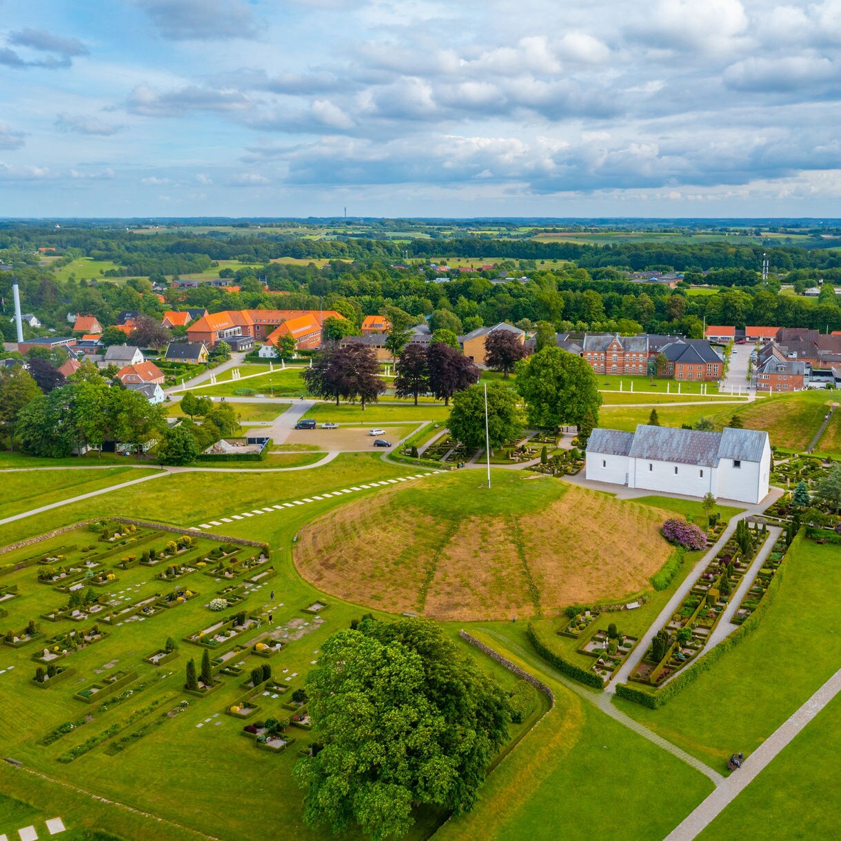 Aerial view of Royal Jelling in Denmark showing white church, burial mounds and surrounding green landscape.