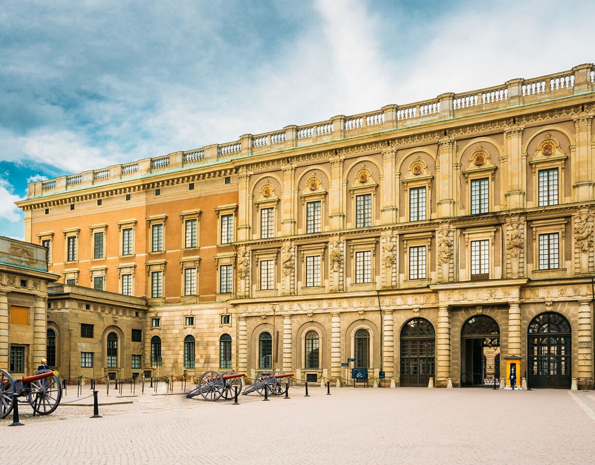 The Royal Palace in Stockholm’s Gamla Stan, featuring ornate stone architecture, arched windows and cannons displayed in the cobblestone courtyard under a cloudy sky.