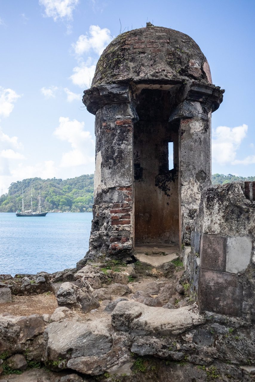 Weathered stone sentry tower at Portobelo ruins in Colón, Panama, overlooking the blue sea with a sailing ship in the distance.