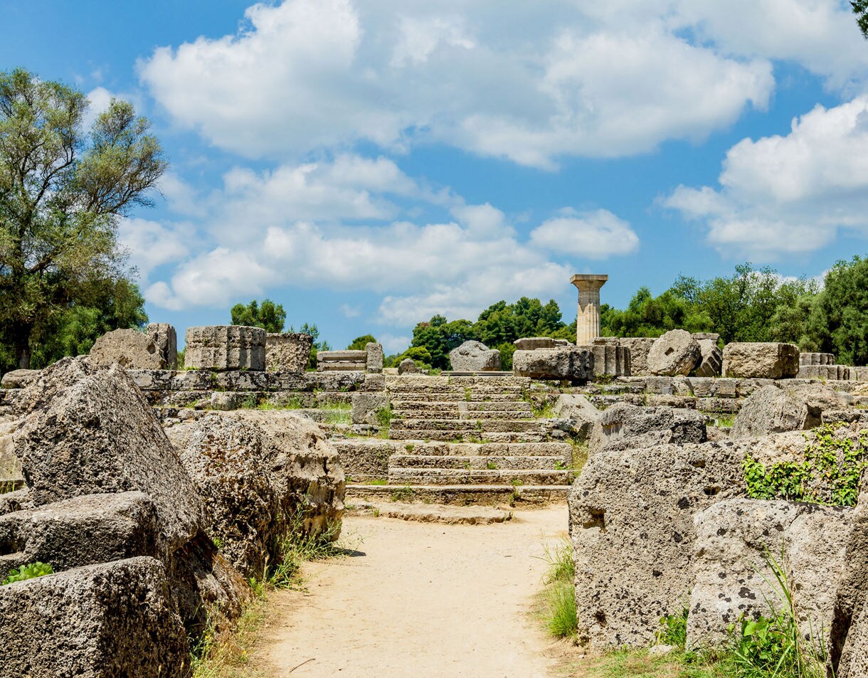 Stone ruins and a lone column at the archaeological site of Olympia, Greece, surrounded by trees and a bright blue sky with scattered clouds.
