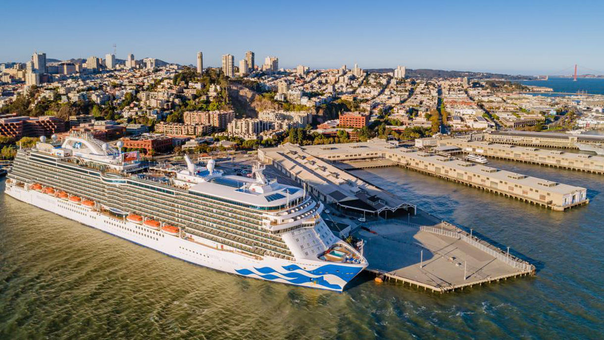 A Princess cruise ship passing under the Golden Gate Bridge in San Francisco, with the city skyline and rolling hills in the background.