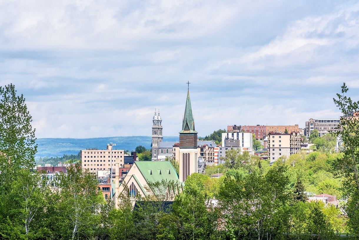 Elevated view of Saguenay, Canada with tall church spires, mid-rise buildings and dense green trees under a bright partly cloudy sky.