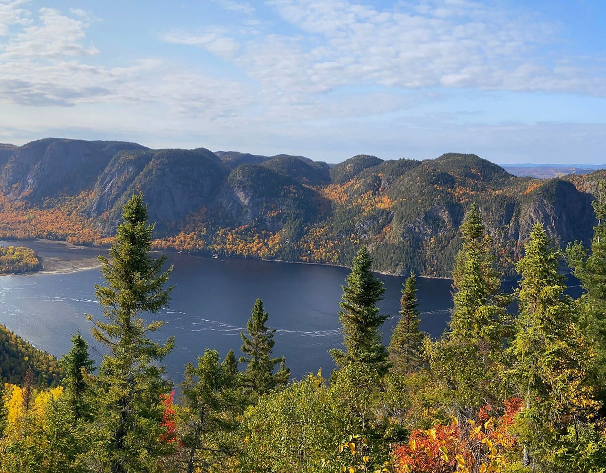 Panoramic view of Saguenay Fjord with dark blue water, steep forested cliffs and vibrant fall foliage seen from a rocky overlook.