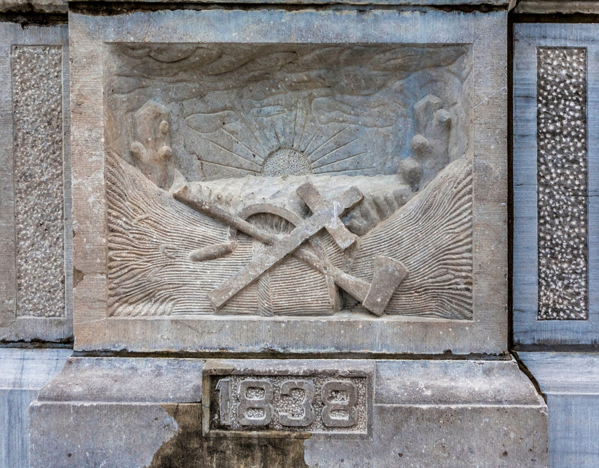A carved stone relief showing a sunburst over hills with crossed tools in the foreground, set above the engraved year 1838.