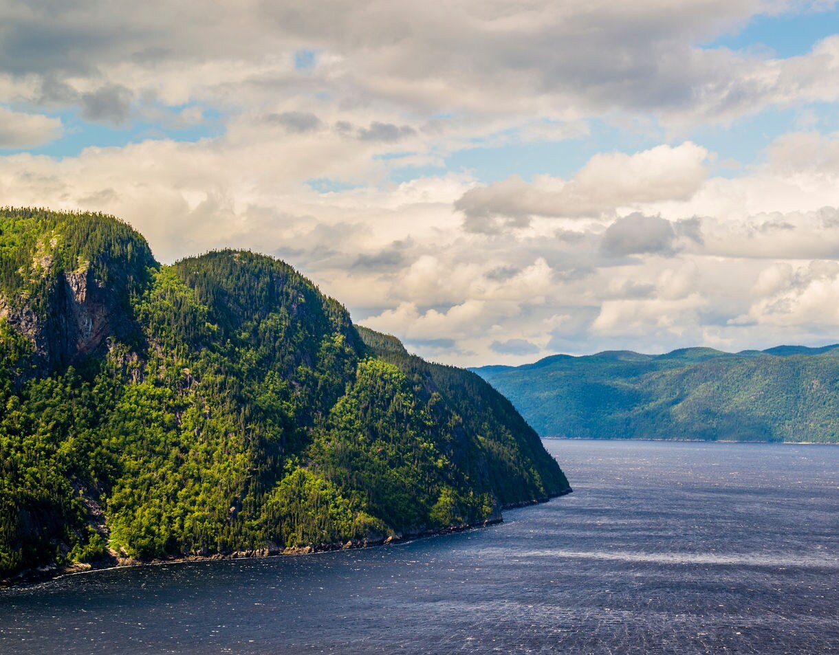 View of Saguenay Fjord with steep forested cliffs on the left, a wide stretch of dark blue water and rolling green hills beneath a sky filled with layered clouds.