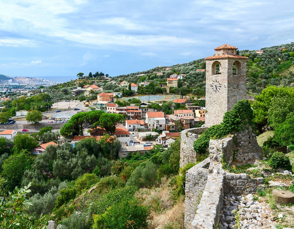 View of Old Bar, Montenegro with a stone clock tower surrounded by lush greenery and clustered houses, with hills and the coastline visible in the distance.