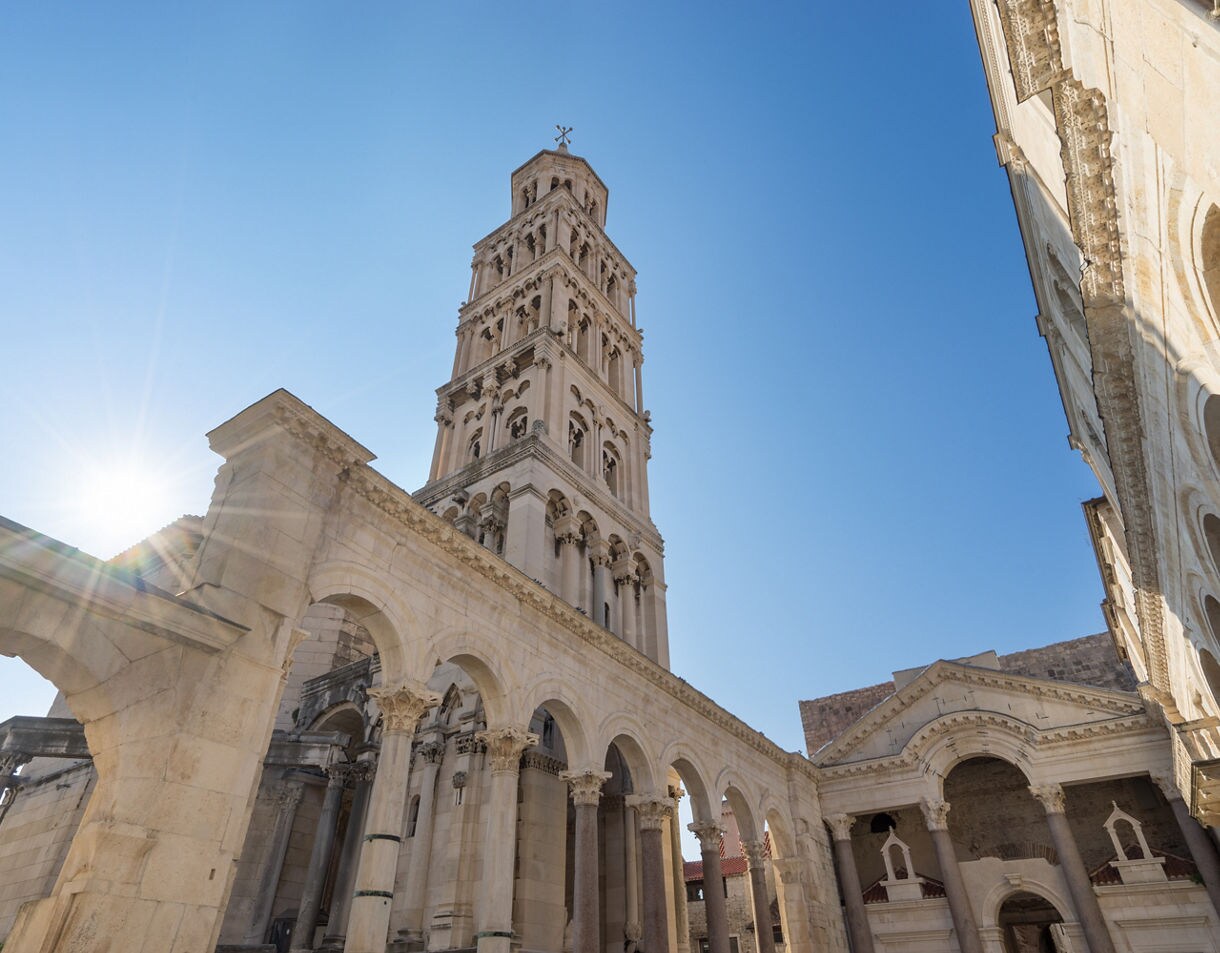Upward view of the bell tower of the Cathedral of Saint Domnius in Split, Croatia, surrounded by Roman arches and pale stone buildings under a bright blue sky with sunlight shining from behind.
