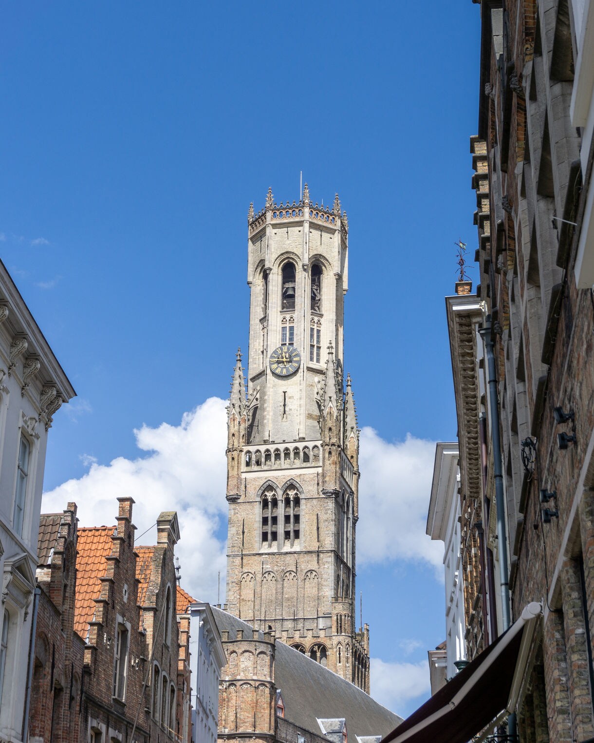 Close-up view of the Belfry of Bruges in Belgium, showing its Gothic clock tower rising above historic brick and stone buildings under a clear blue sky.
