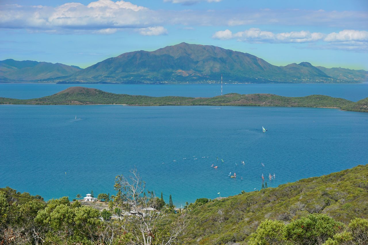 Elevated view of Sainte-Marie’s turquoise lagoon dotted with colorful windsurfers, with green hills and a tall mountain range rising in the background.