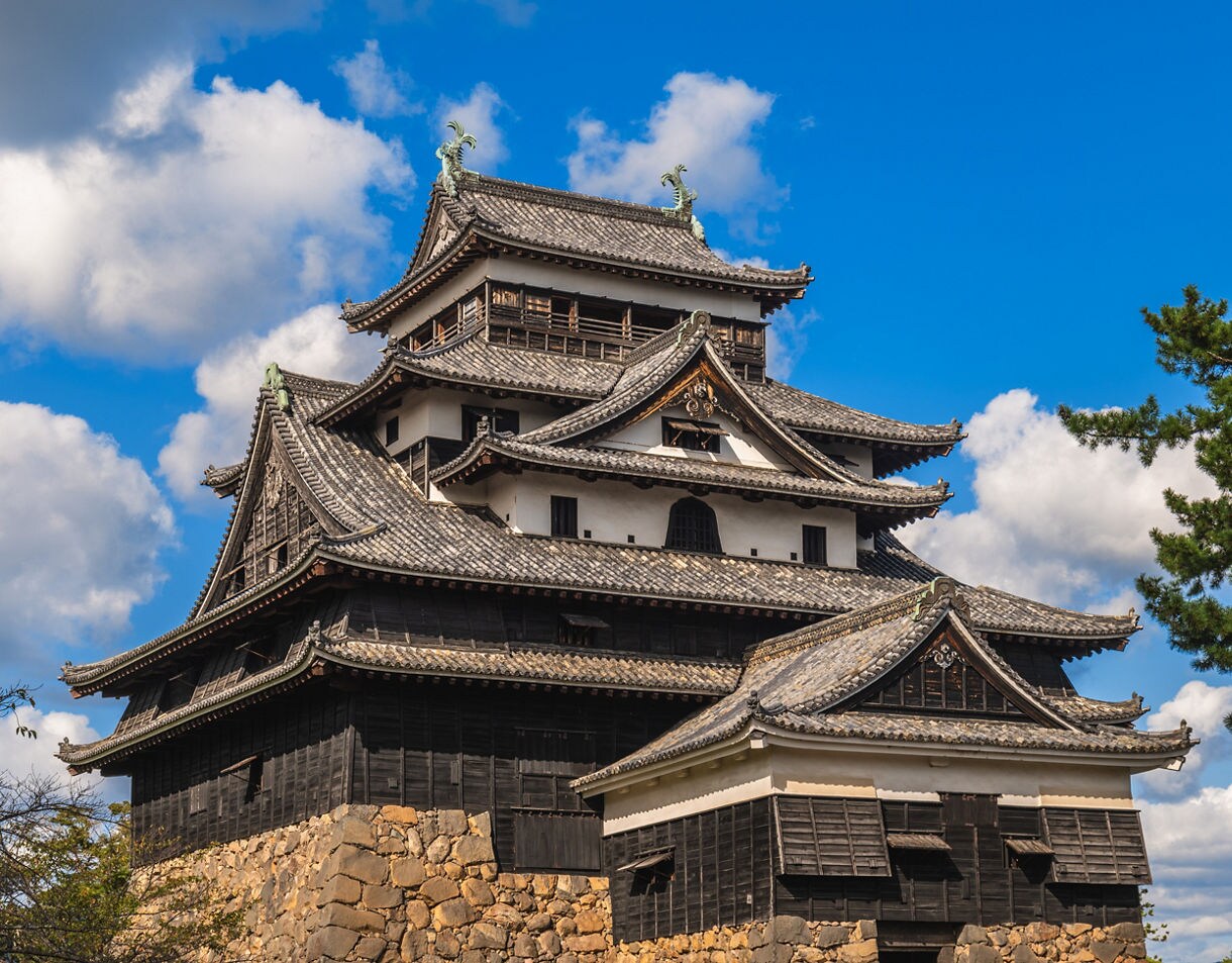Dark wooden Japanese castle with layered tile roofs and stone base, set against a blue sky with puffy clouds.