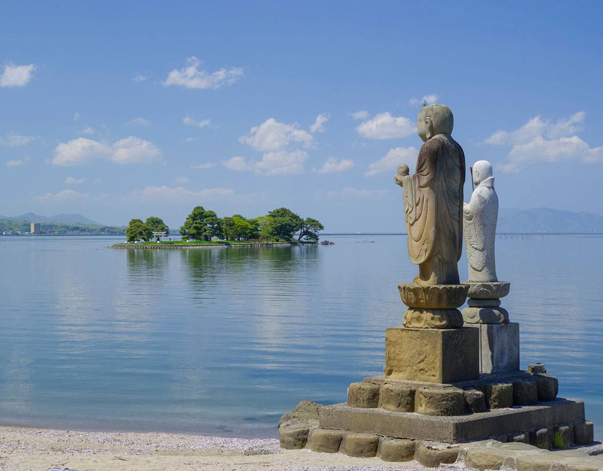 Stone statues on the lakeshore facing a small tree-covered island with a white torii gate in the calm waters of Lake Shinji.