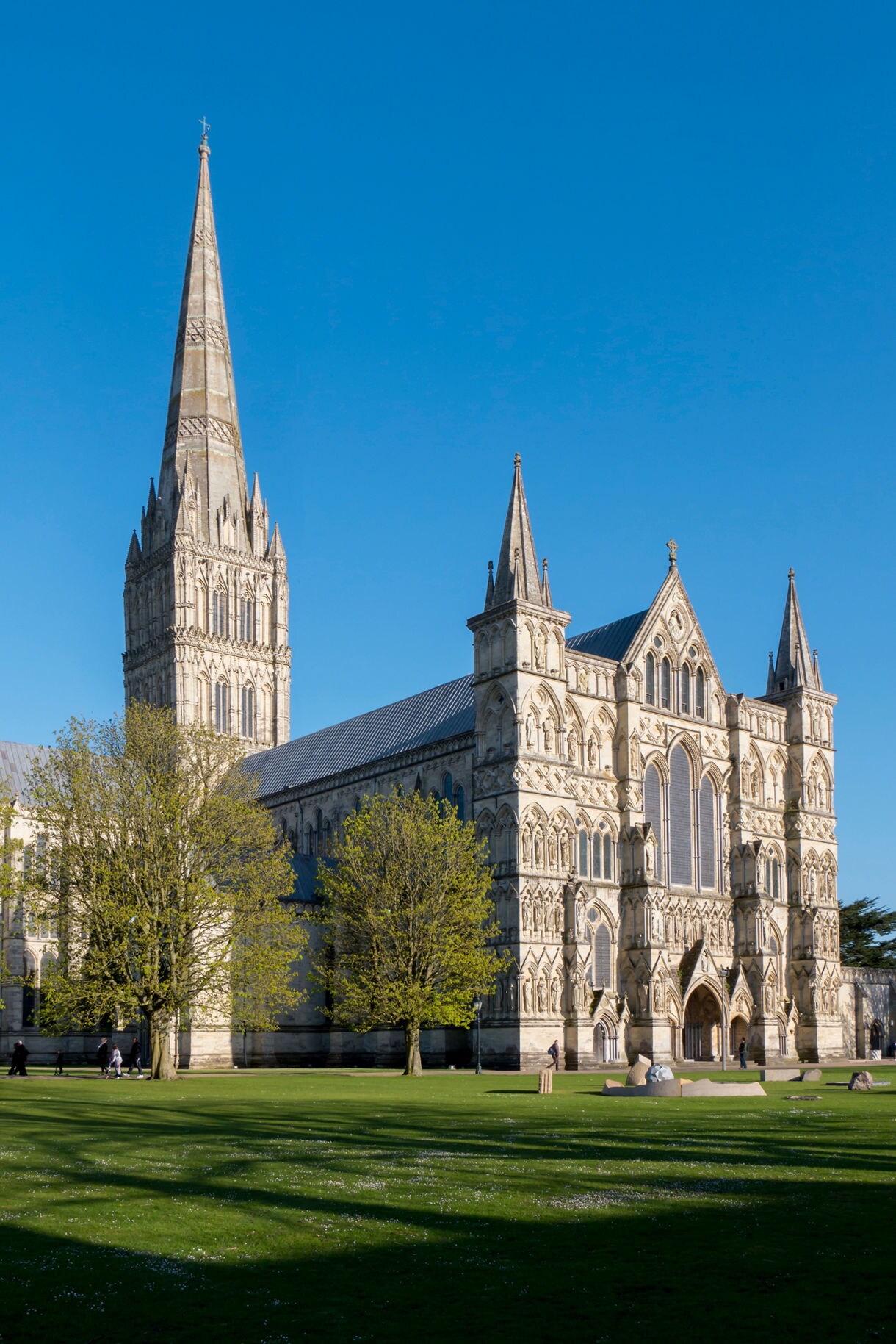 Vertical view of Salisbury Cathedral in Wiltshire, showing its tall Gothic spire and ornate façade framed by green lawns and trees under a bright blue sky.