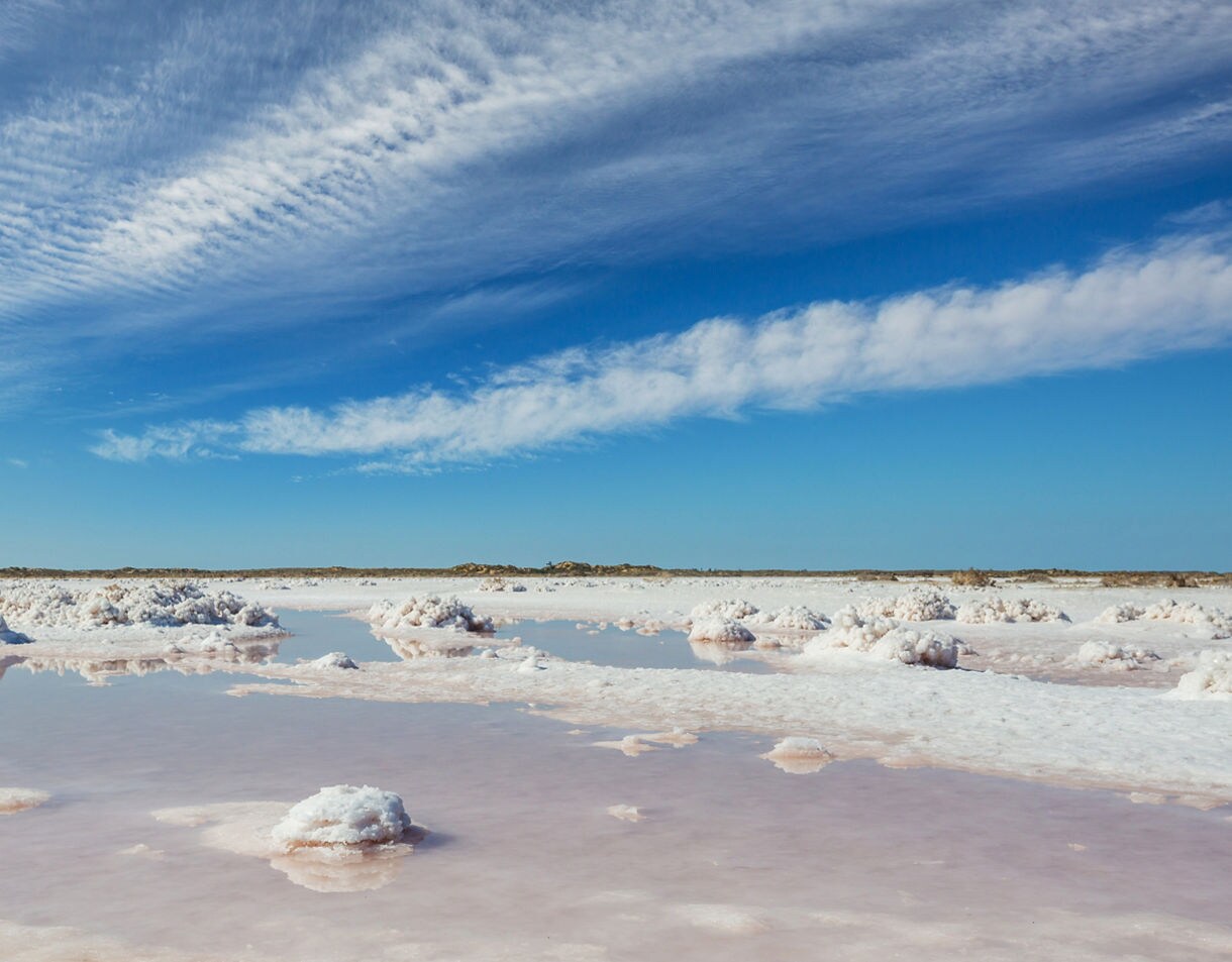 Expansive salt flats with pinkish water pools and white salt mounds beneath a bright blue sky streaked with clouds.