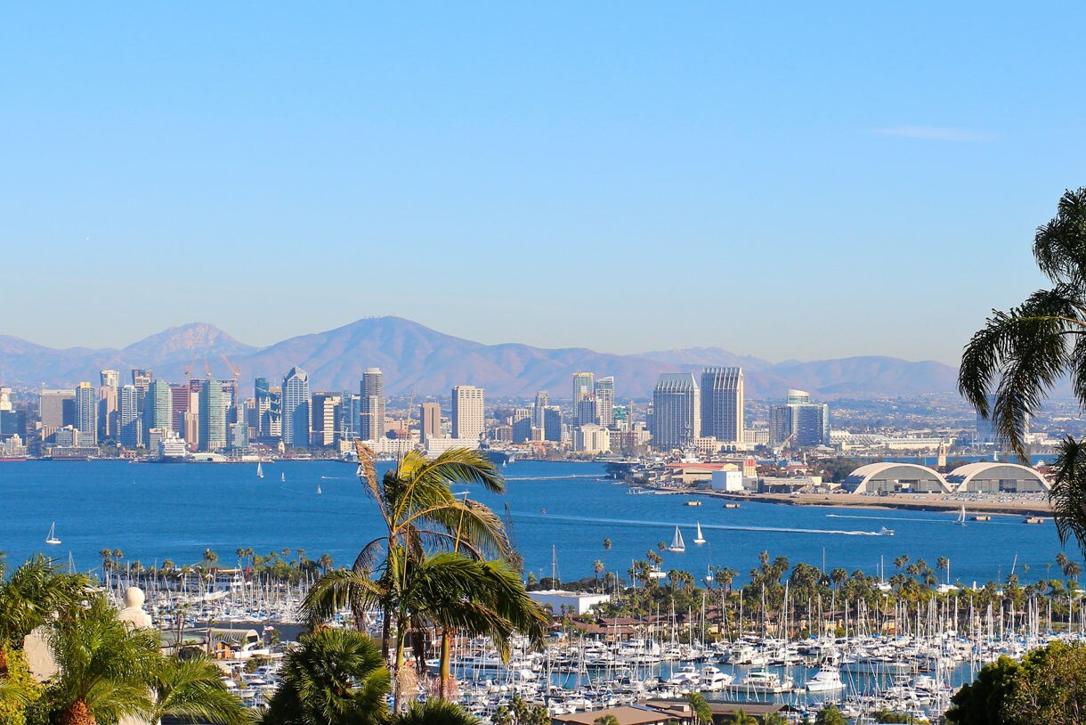 View of San Diego’s downtown skyline with tall buildings, a busy marina, and surrounding mountains under a clear blue sky.