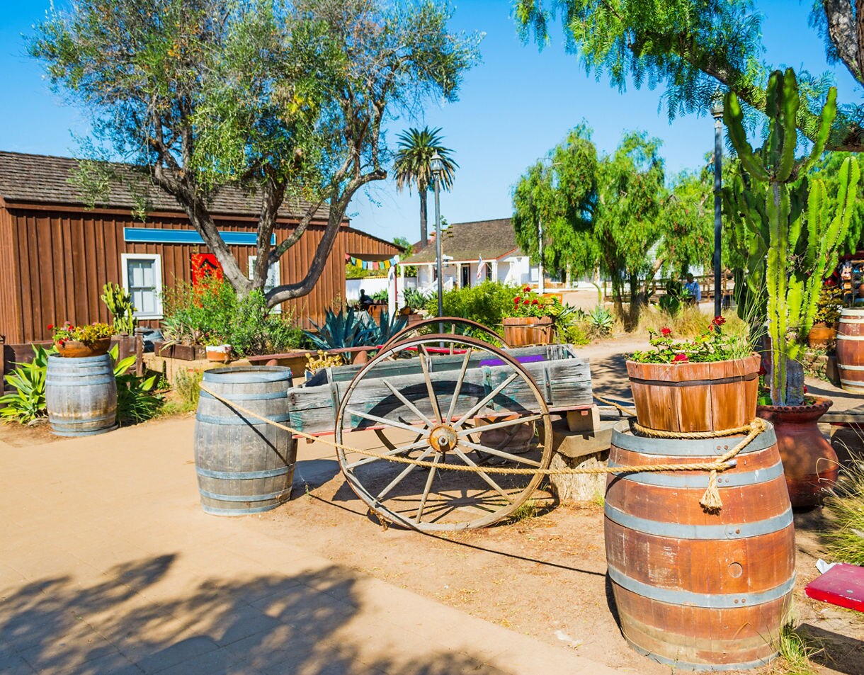 Wooden wagon and barrels decorated with flowers in San Diego’s Old Town surrounded by trees, cacti, and historic wooden buildings.