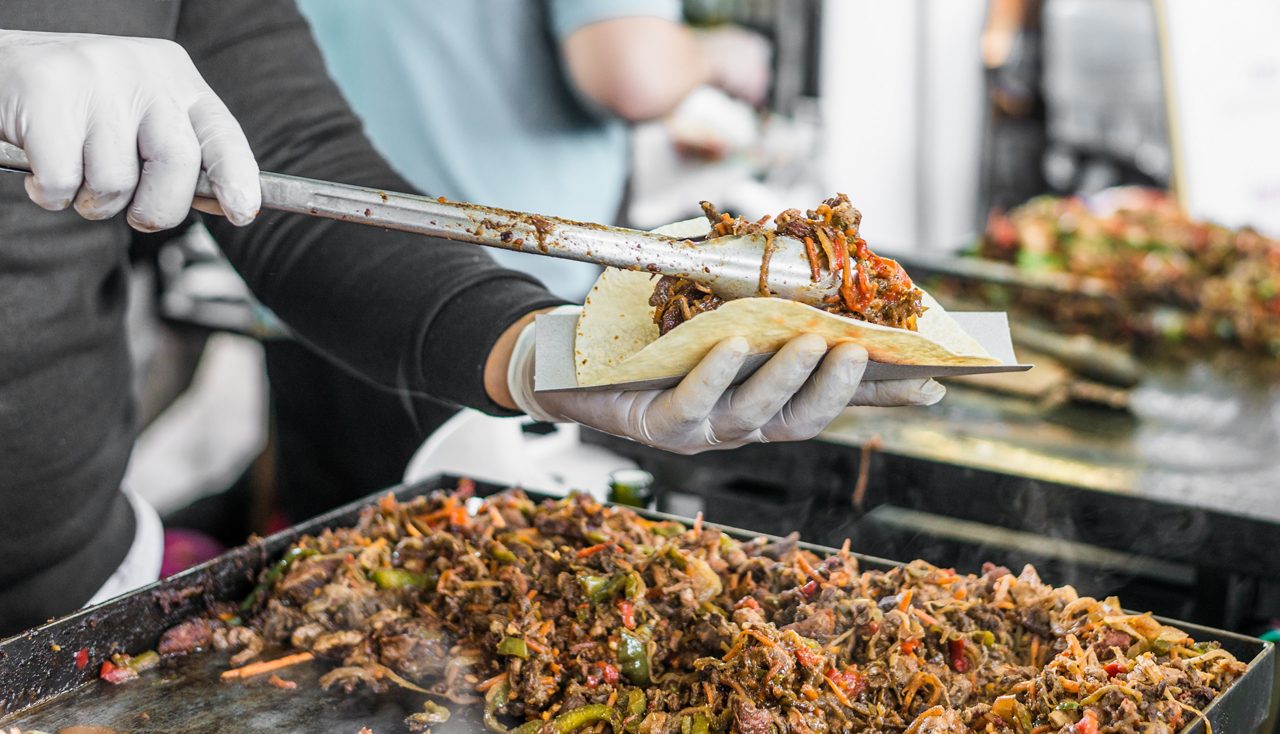 Close-up of a vendor preparing a taco with grilled meat and vegetables using tongs at a food stand in San Diego.