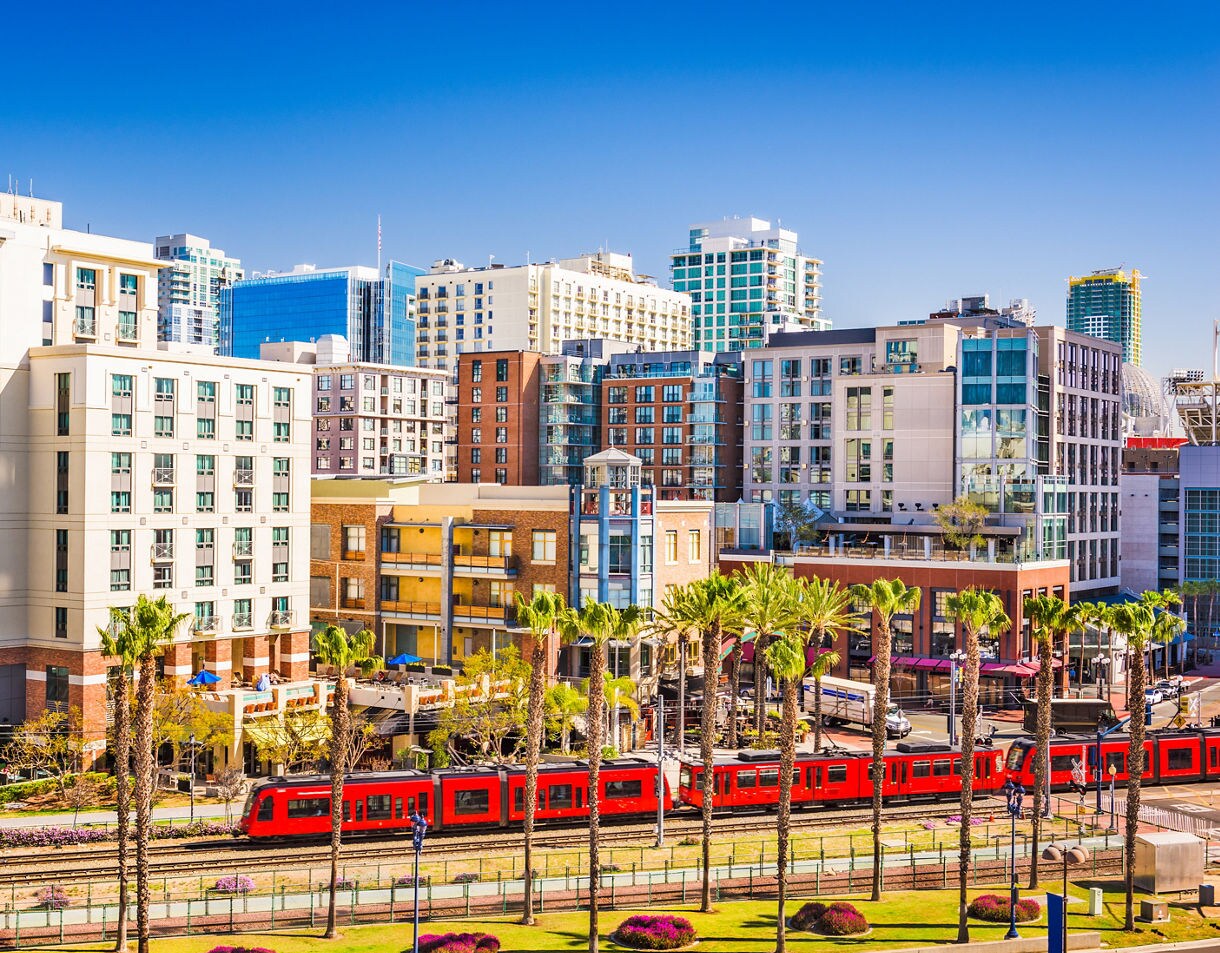 Red San Diego trolley passing through downtown with modern buildings, palm trees, and clear blue skies in the background.