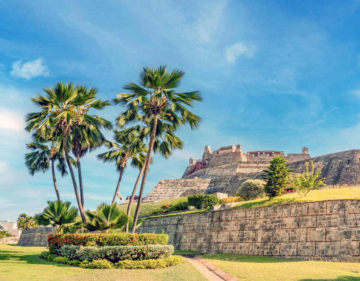 Stone walls of San Felipe Fortress in Cartagena, Colombia with palm trees and manicured gardens under a bright blue sky.