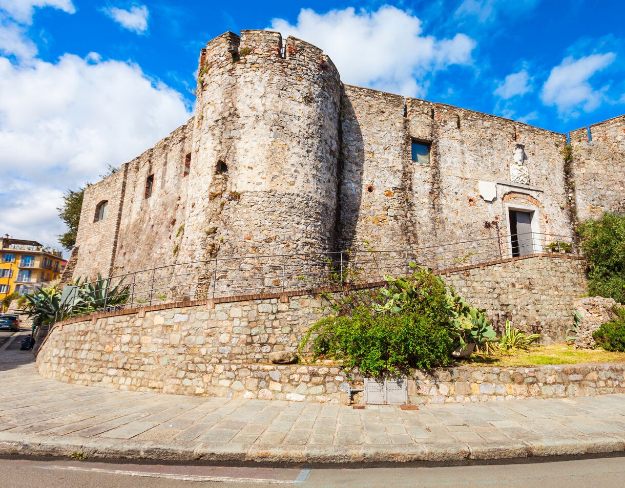 A fortress built of rough gray stone with rounded towers and small windows, sitting on a raised stone terrace with plants and a paved walkway in front under a bright blue sky.