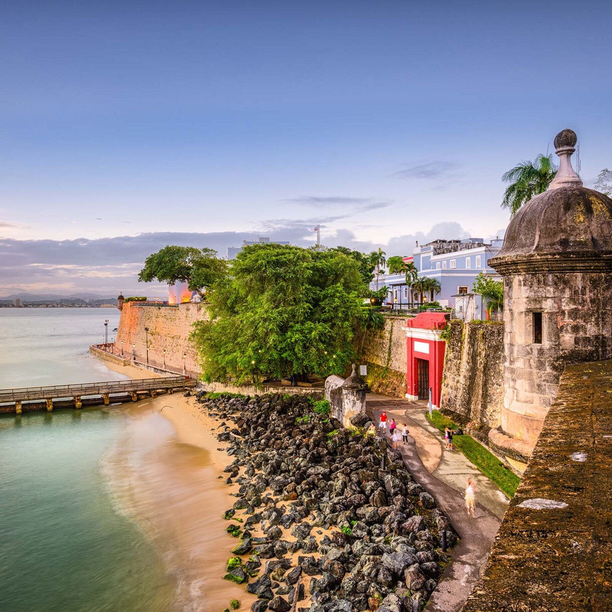 The historic San Juan Gate in Puerto Rico, framed by ancient stone walls, lush greenery, and a scenic coastline with calm waters at sunset.